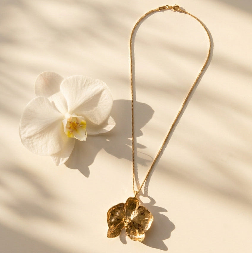 A gold necklace with a textured, orchid shaped pendant, displayed on a light gray textured background.