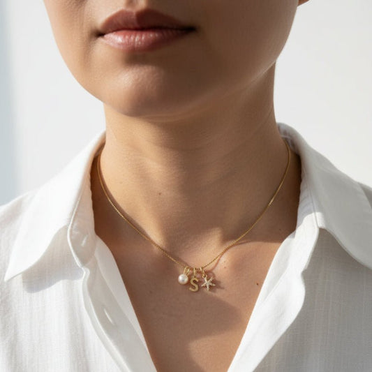 A close-up of a person's neck and upper chest wearing a delicate gold necklace with a small pendant featuring a white pearl and gold lettering, paired with a black shirt.