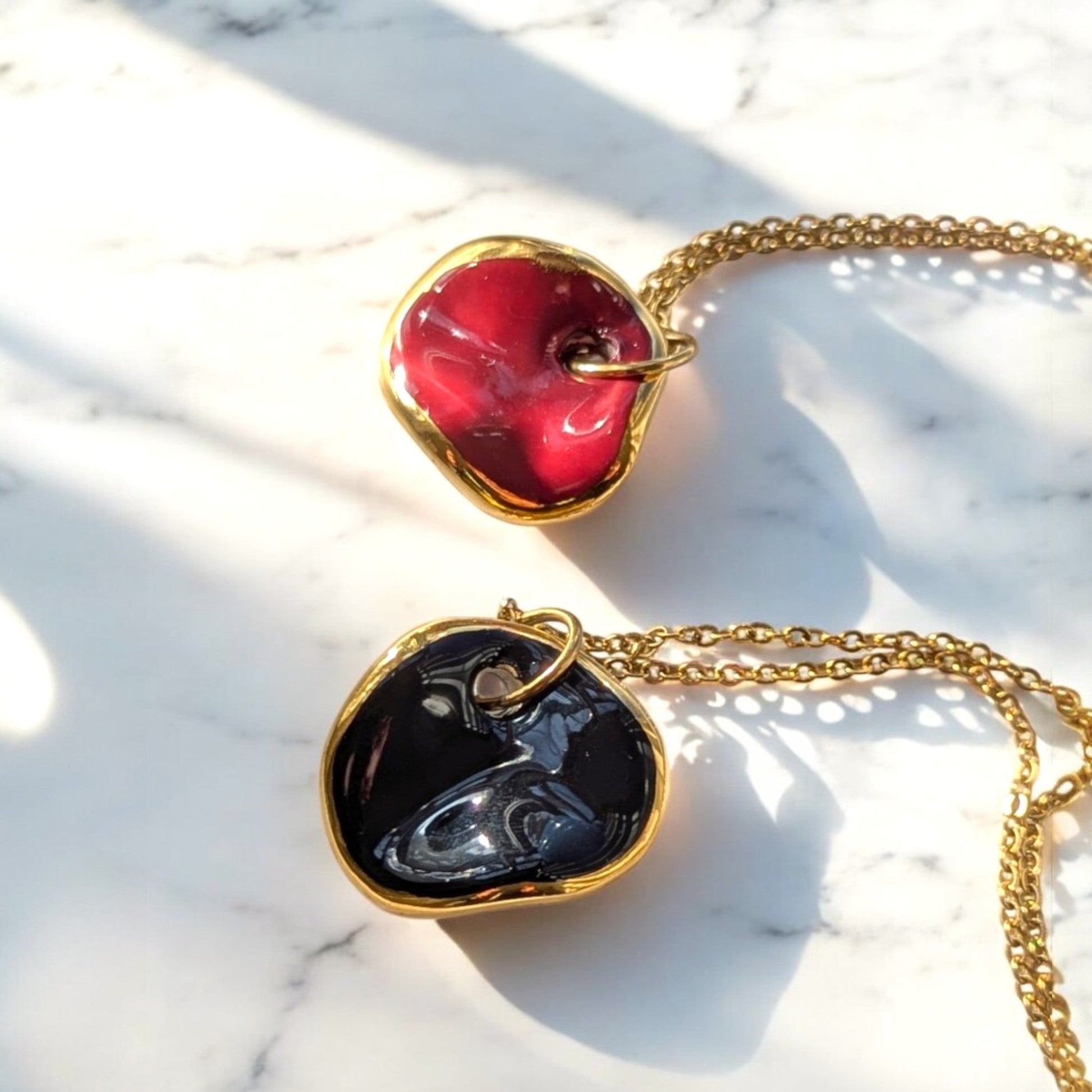 Two Maximilianas Ondulado Moyen ceramic pendant necklaces , one featuring a red ceramic pendant and the other with a black ceramic pendant, both on a gold chain, displayed on a marble surface - Close up shot.