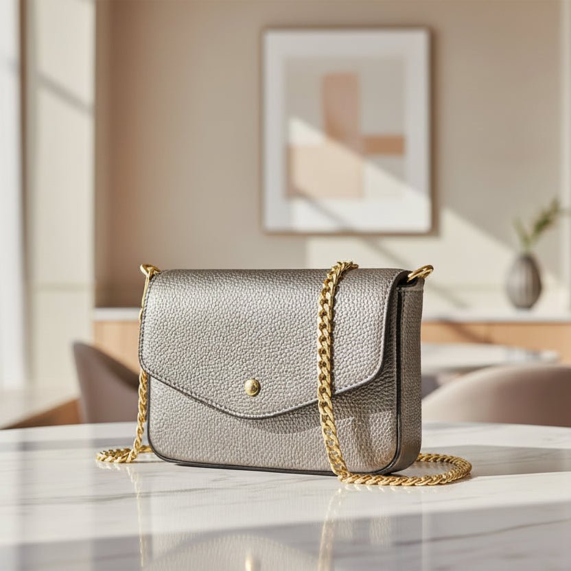 A metallic taupe pebbled leather crossbody bag with a gold chain strap and a small gold button closure, shown on a table in a chic restaurant